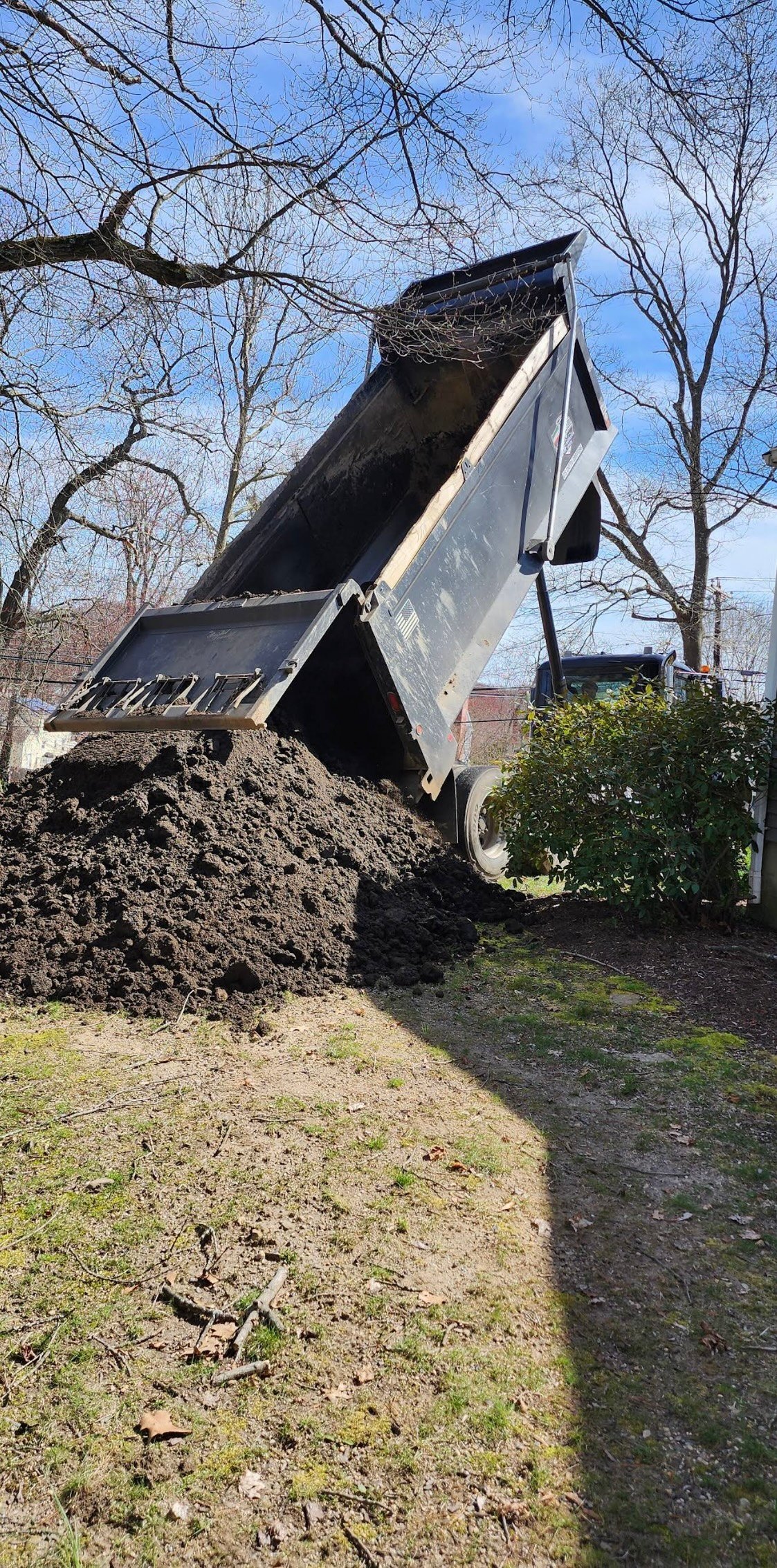 Dump truck unloading dark soil or mulch onto a residential yard with bare trees in background