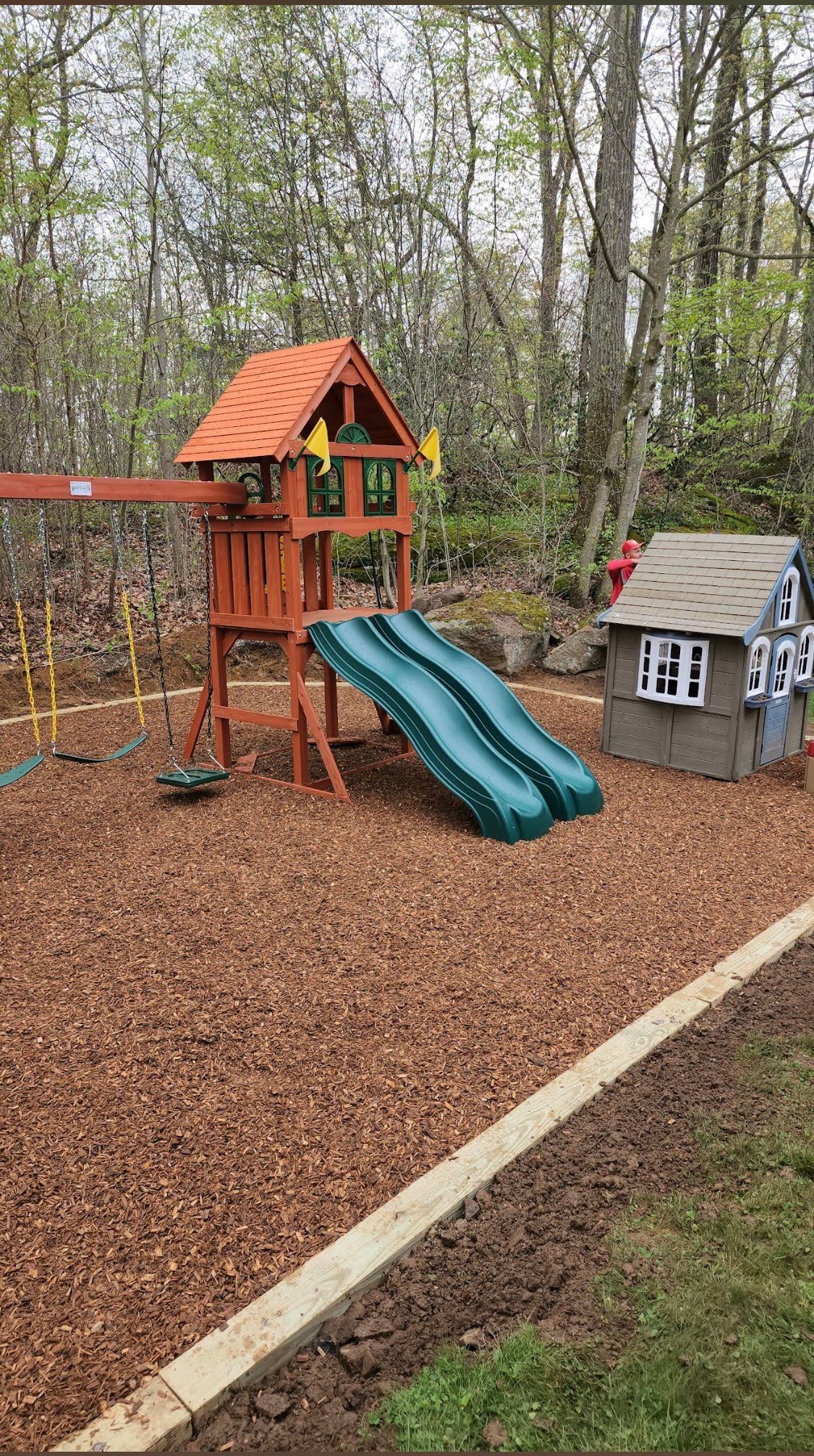 Backyard playground with red wooden play structure featuring a green slide, small playhouse, and swings surrounded by mulch and tall trees