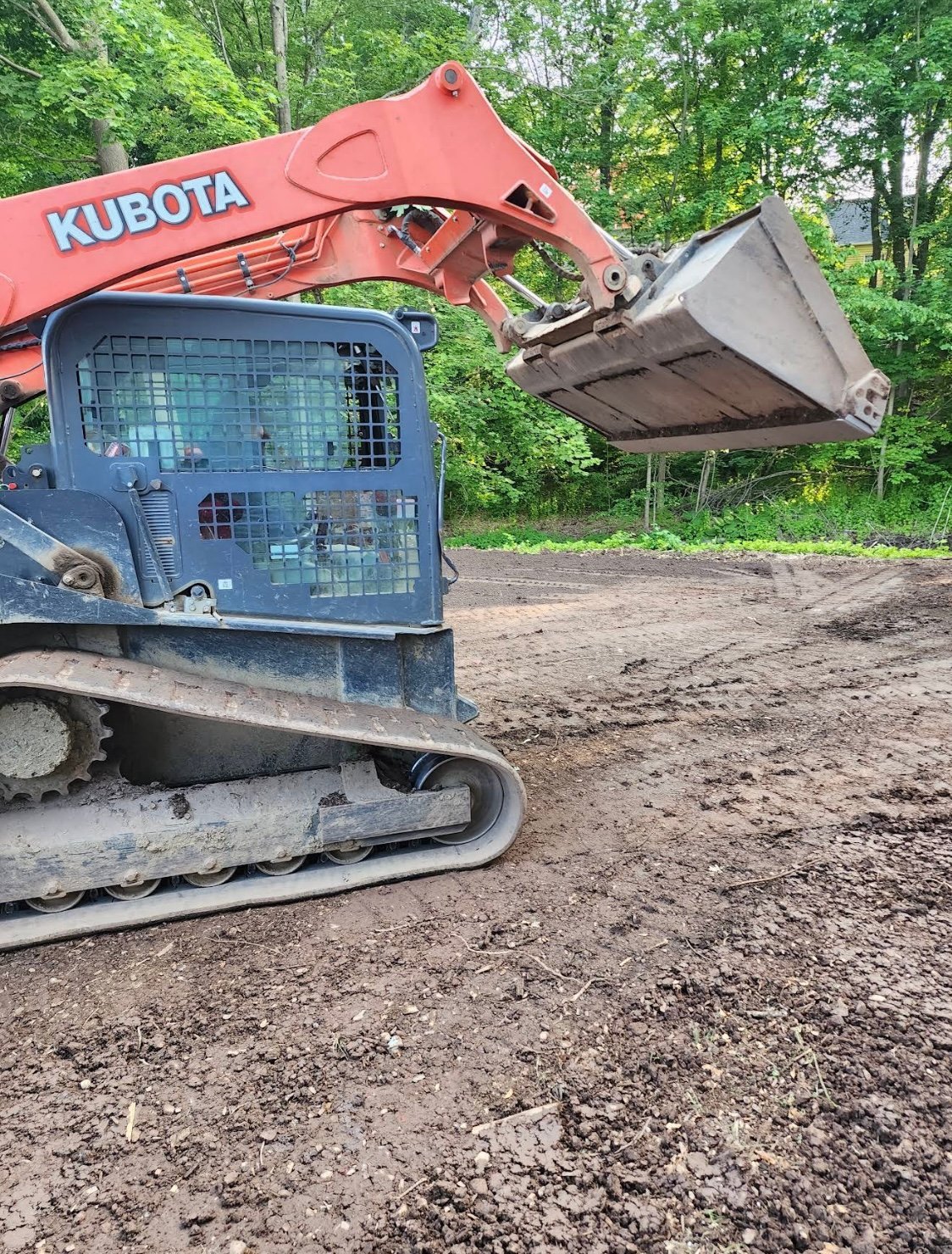 Red and blue Kubota mini excavator with bucket attachment on dirt construction site surrounded by trees