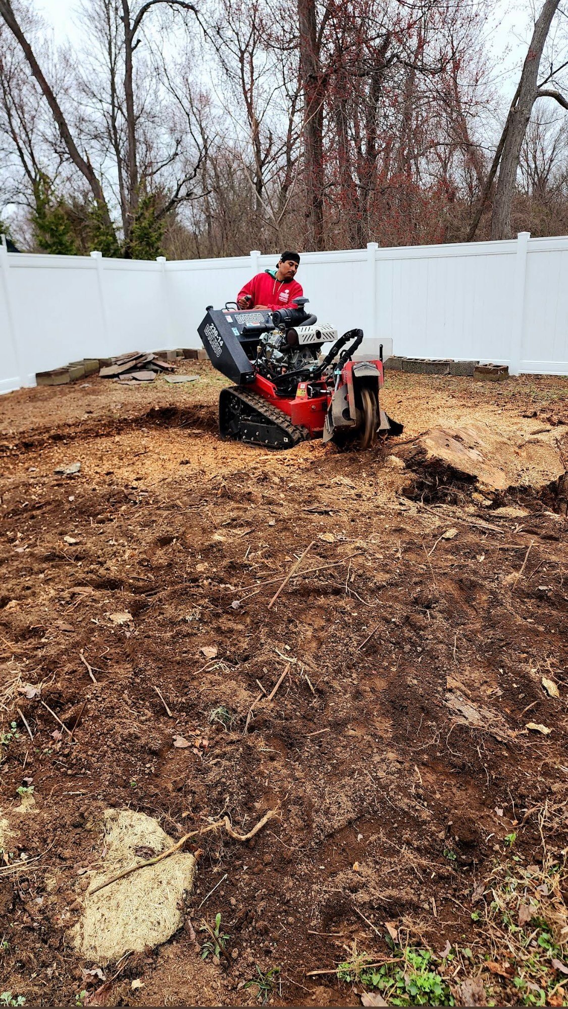Person operating a red and black tracked loader in a dirt yard with bare trees and white fence in background