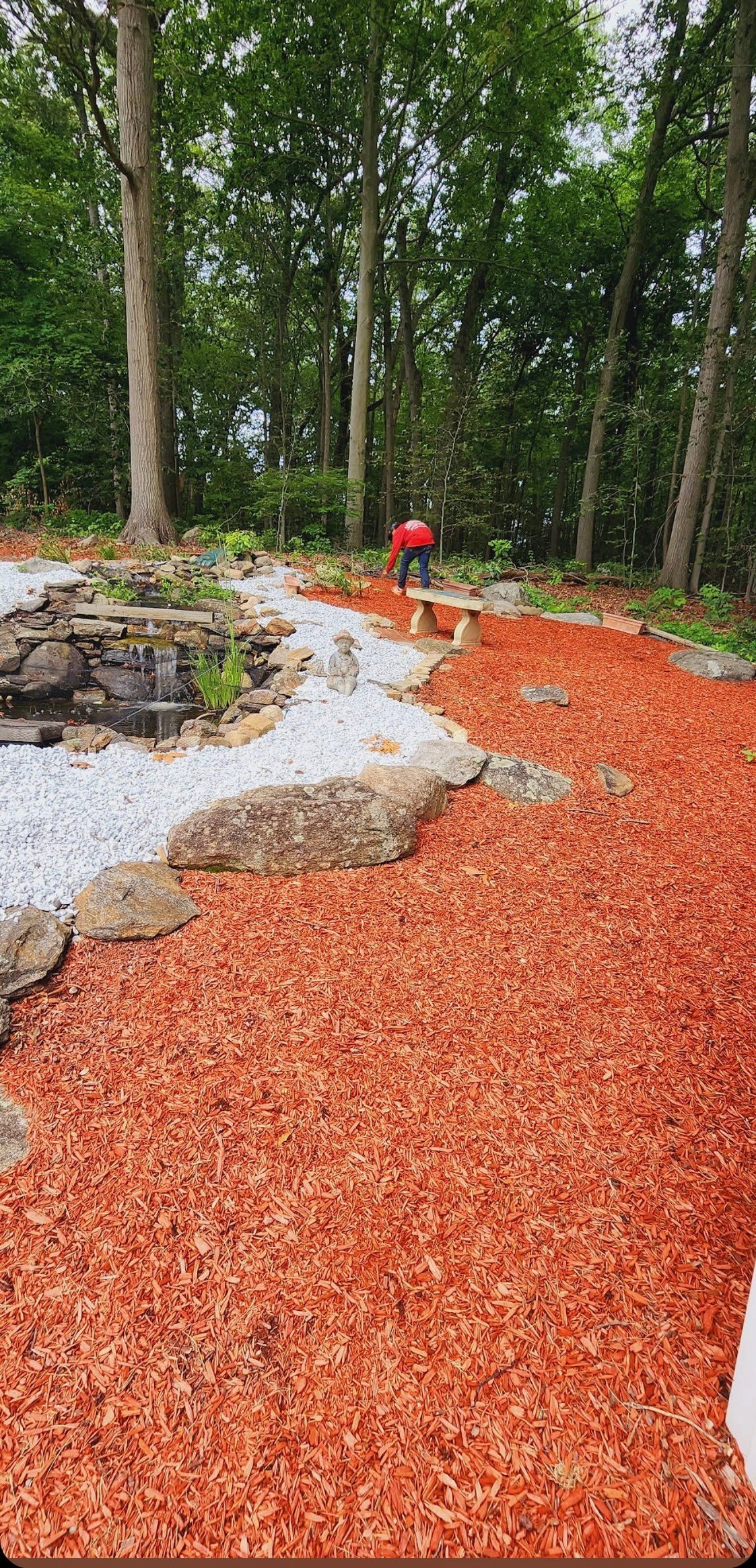 Forested nature playground with red mulch ground, white gravel paths, rocks, a person in red, and tall trees