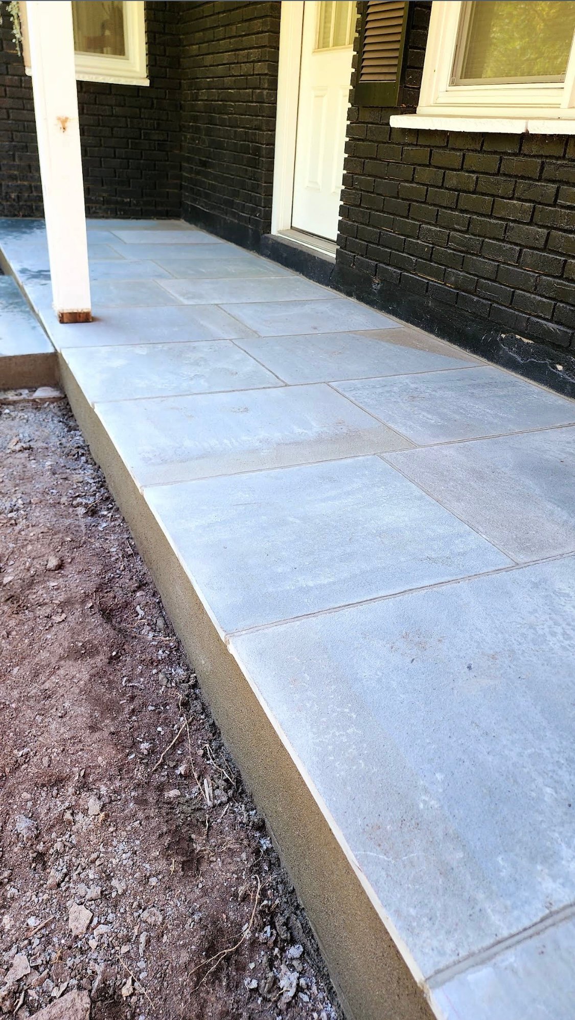Newly installed light blue-gray patio pavers on a concrete foundation against a black brick building with white trim and windows