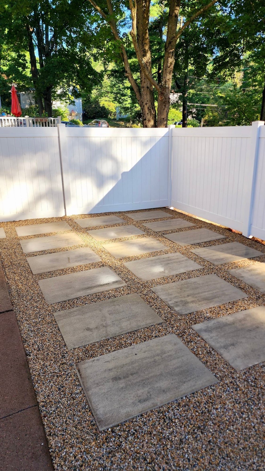 Backyard patio with concrete pavers arranged in grid pattern surrounded by gravel, bordered by white vinyl fence and tall green trees