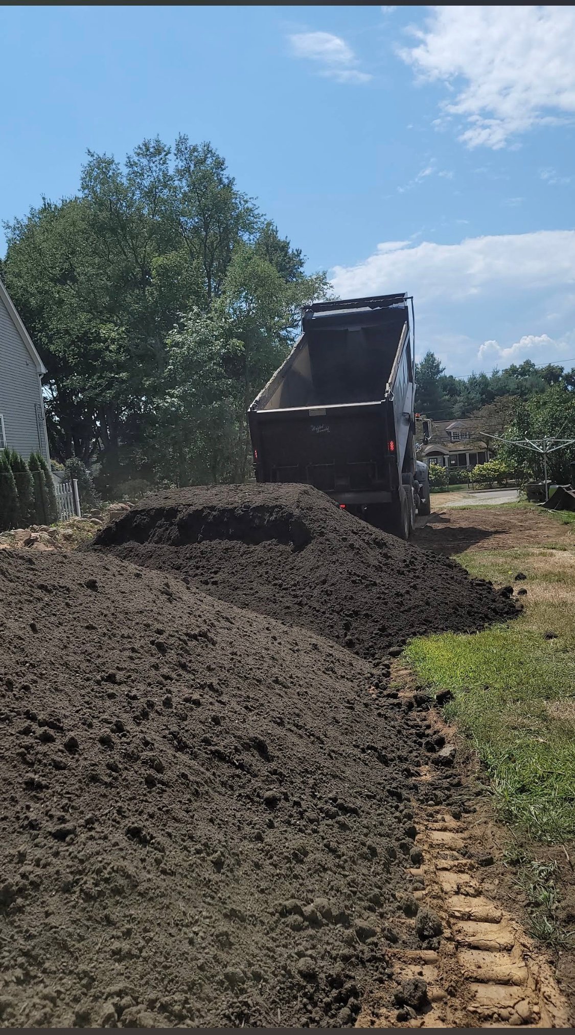 Dump truck unloading soil onto a residential driveway with trees and house in background