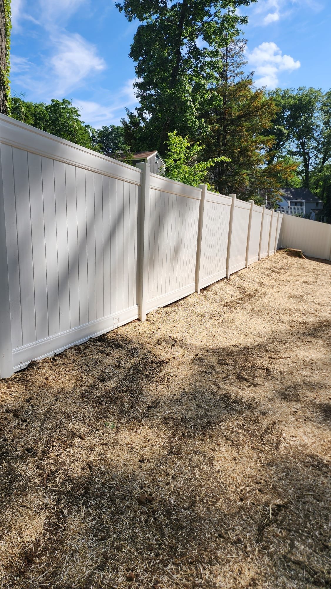 Tall white vinyl privacy fence panels installed along a cleared dirt path with trees and blue sky in the background