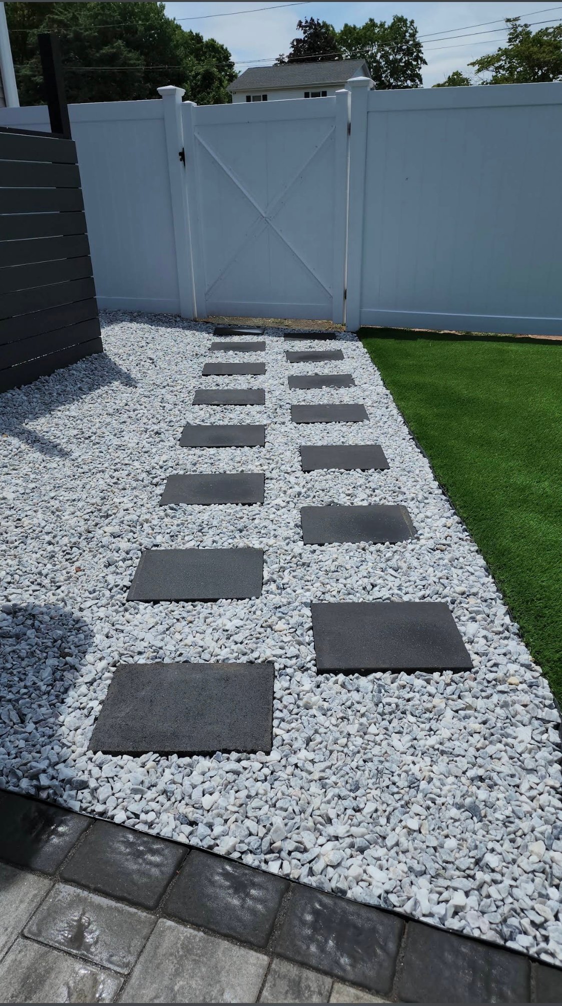 Backyard pathway with dark rectangular stepping stones set in white pea gravel between green artificial turf and white vinyl fencing