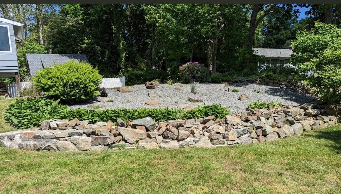 Backyard landscape with a curved rock border edging a gravel garden bed, surrounded by green trees and grass