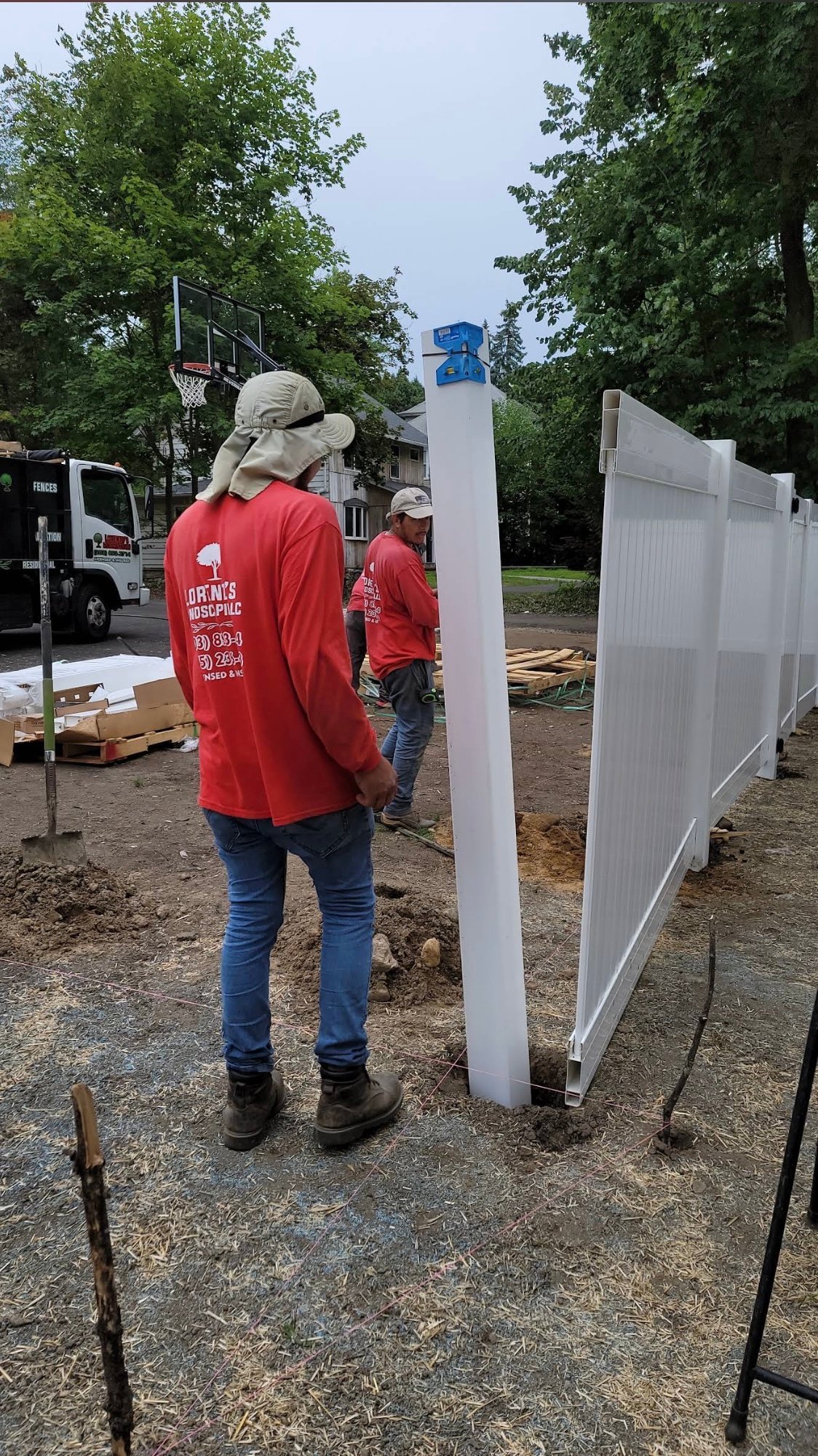 Workers in red shirts assembling white fencing panels at an outdoor construction site with trees and equipment in background