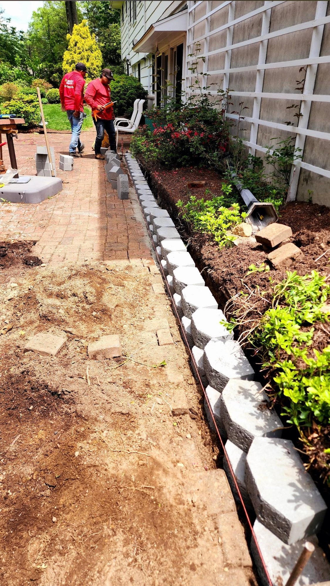Two people in red jackets work on a brick patio beside a house with newly installed white garden edging and mulched flower beds