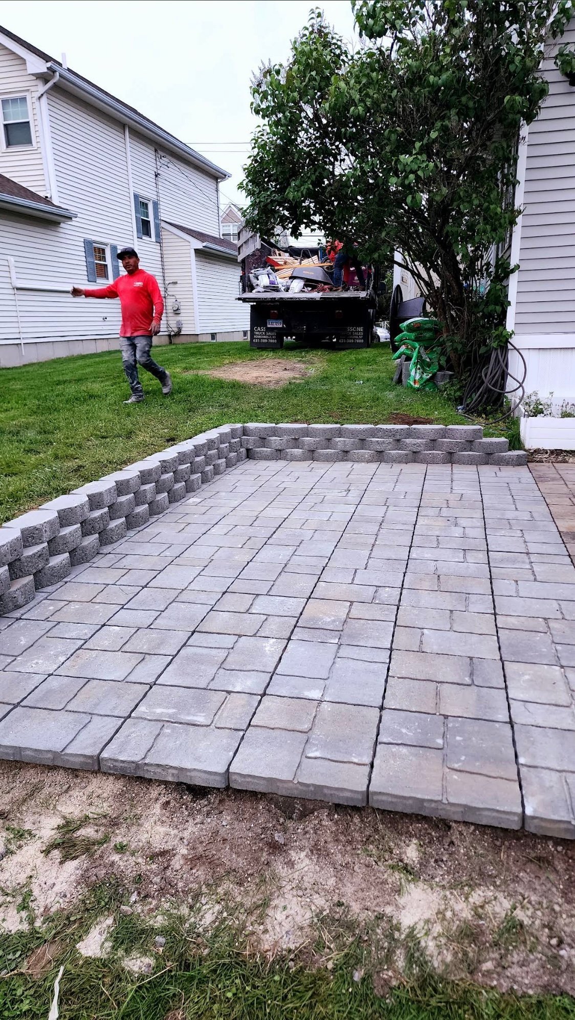 New gray paver patio in residential backyard with brick retaining wall, landscaping truck in background, and houses on either side