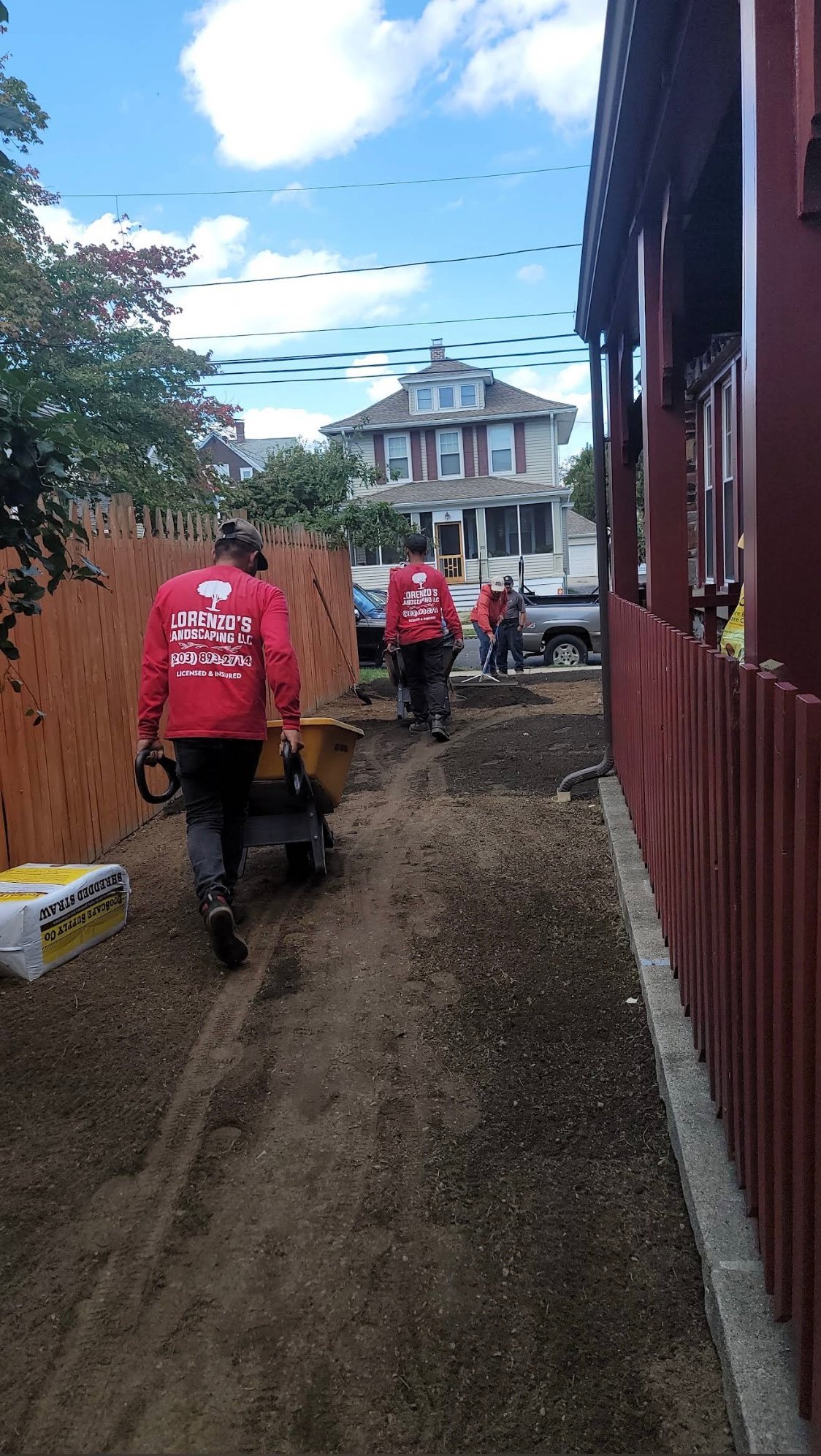 Construction workers in red shirts work on a residential alley with wooden fences and a colonial-style house visible in the background