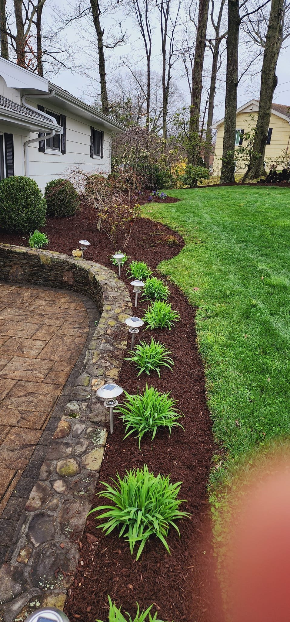 Landscaped garden bed with green plants and solar lights along a stone retaining wall beside a brick patio
